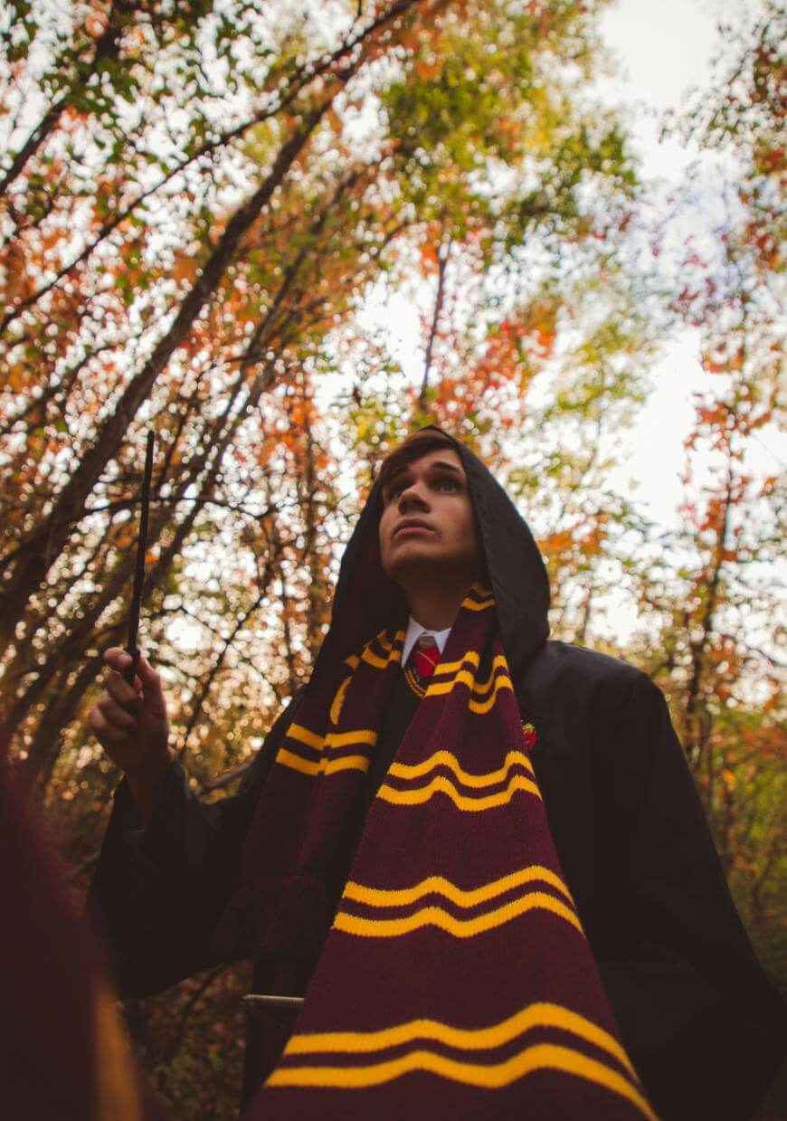 A person wearing a black cloak and a maroon and yellow striped scarf holds a wand while standing in a forest with autumn trees, looking upwards.