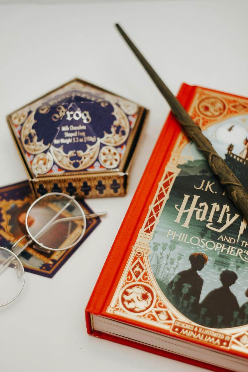 A Harry Potter book, a wand, round glasses, a chocolate frog box, and a card are arranged on a white surface.