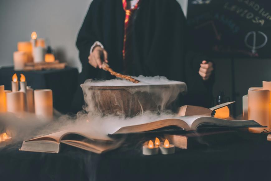 A person in a robe with a striped tie stirs a smoking cauldron surrounded by open books and glowing candles, creating a magical or wizard-like atmosphere.