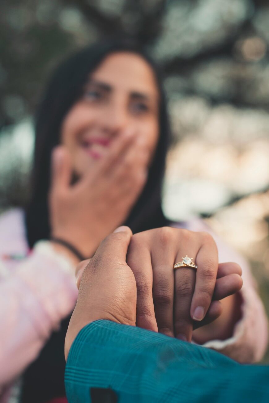 A smiling woman covers her mouth in surprise while showing off an engagement ring, as another person gently holds her hand. The background is softly blurred.