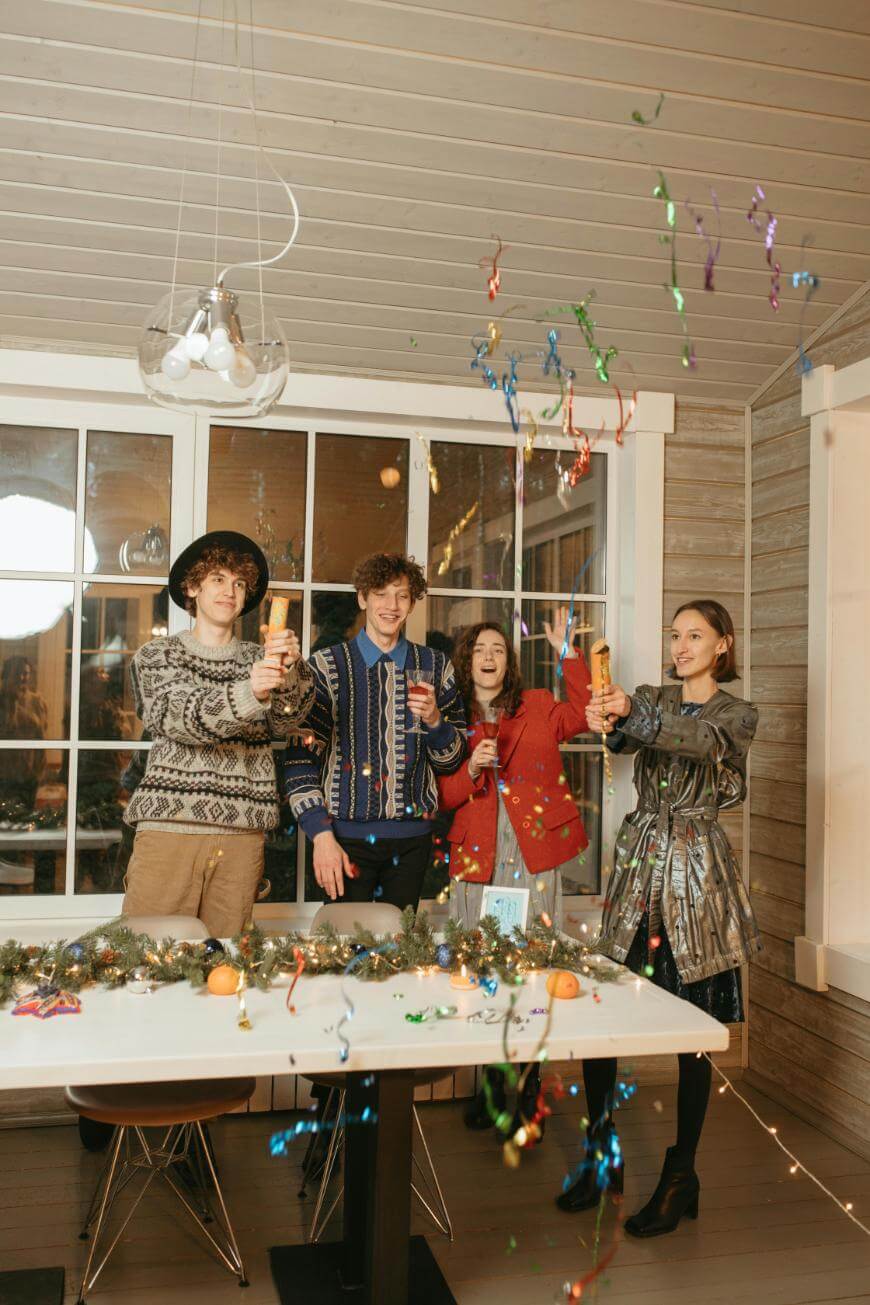 Four people in festive outfits stand behind a decorated table, smiling and launching colorful confetti poppers in a warmly lit room with holiday garlands and ornaments.