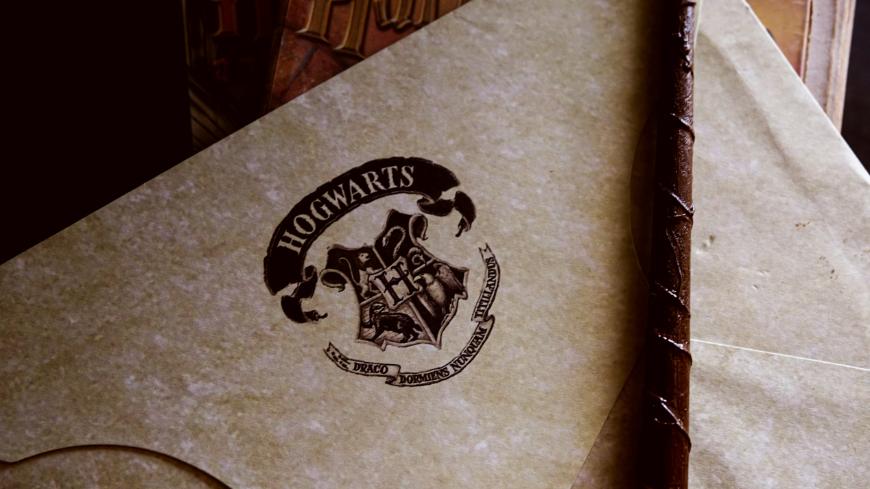 A close-up of a parchment envelope with the Hogwarts crest and motto printed on it, next to a wand, evoking the magical world of Harry Potter.