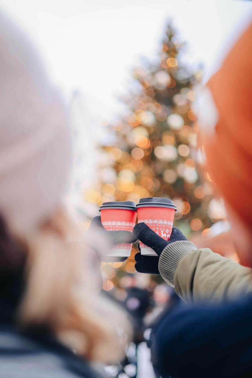 Two people in winter hats and gloves hold up festive red coffee cups in front of a blurred, decorated Christmas tree with lights in the background.
