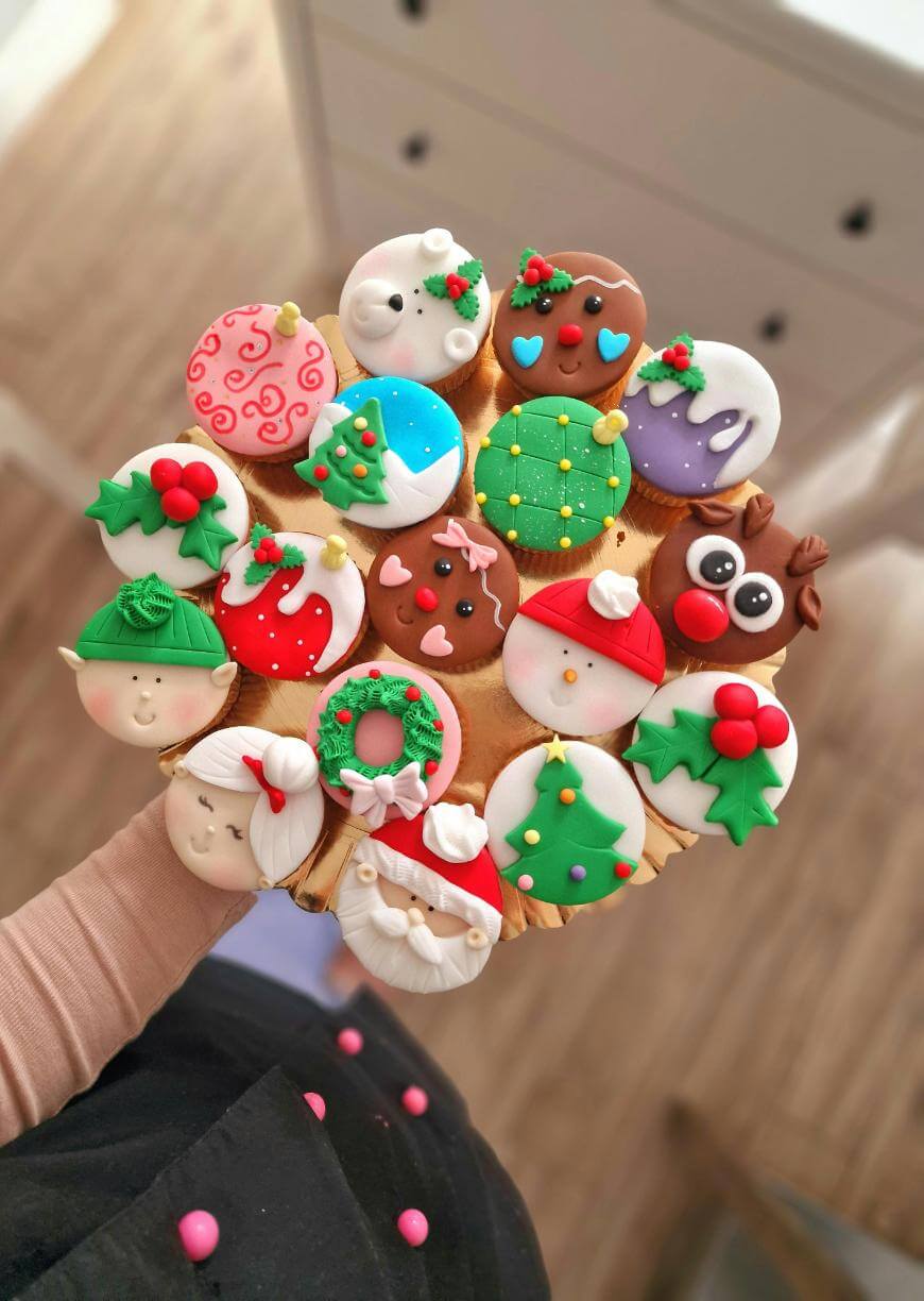 A hand holds a round wooden tray with 15 colorful Christmas-themed cupcakes, decorated with fondant faces, wreaths, ornaments, Santa, reindeer, elves, holly, and festive patterns.