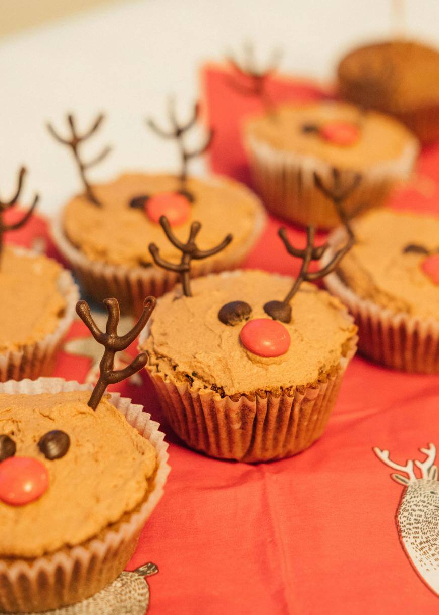 Chocolate cupcakes decorated as reindeer with pretzel antlers, red candy noses, and chocolate chip eyes, arranged on a red napkin with a reindeer illustration.