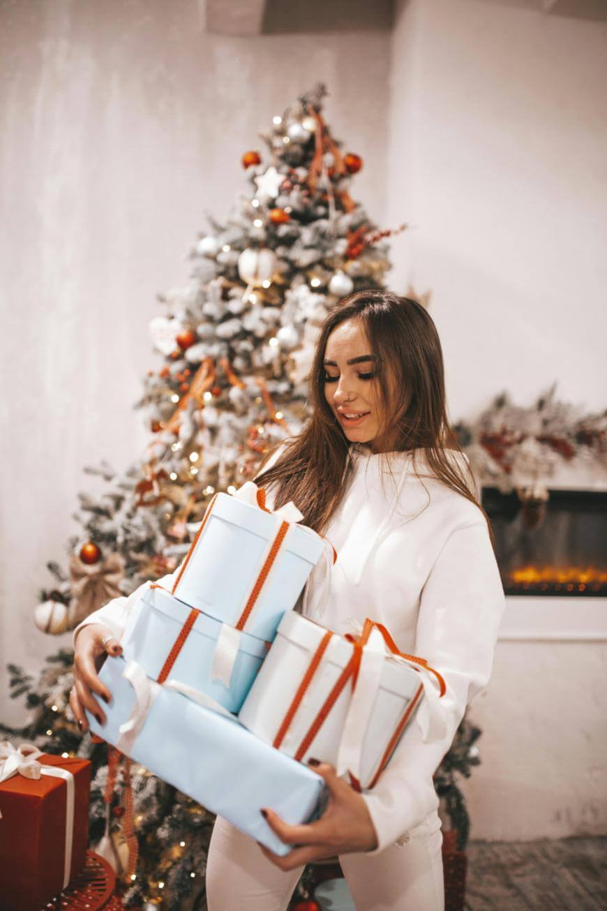 A woman in white clothing holds several wrapped gifts with red ribbons, standing in front of a decorated Christmas tree and a fireplace in a festive, cozy room.