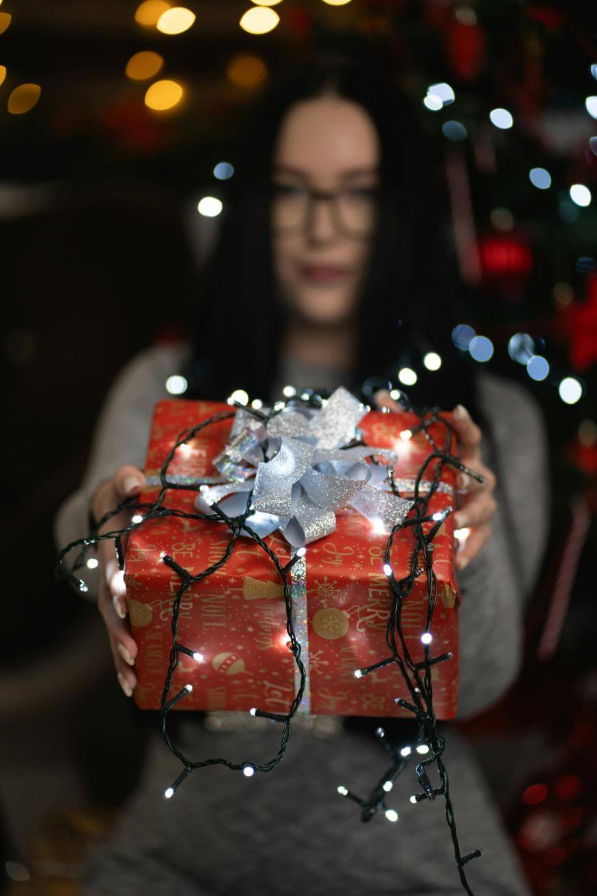 A woman with long dark hair and glasses holds out a red, gift-wrapped present decorated with a silver bow and string lights. Blurred holiday lights and a Christmas tree are visible in the background.