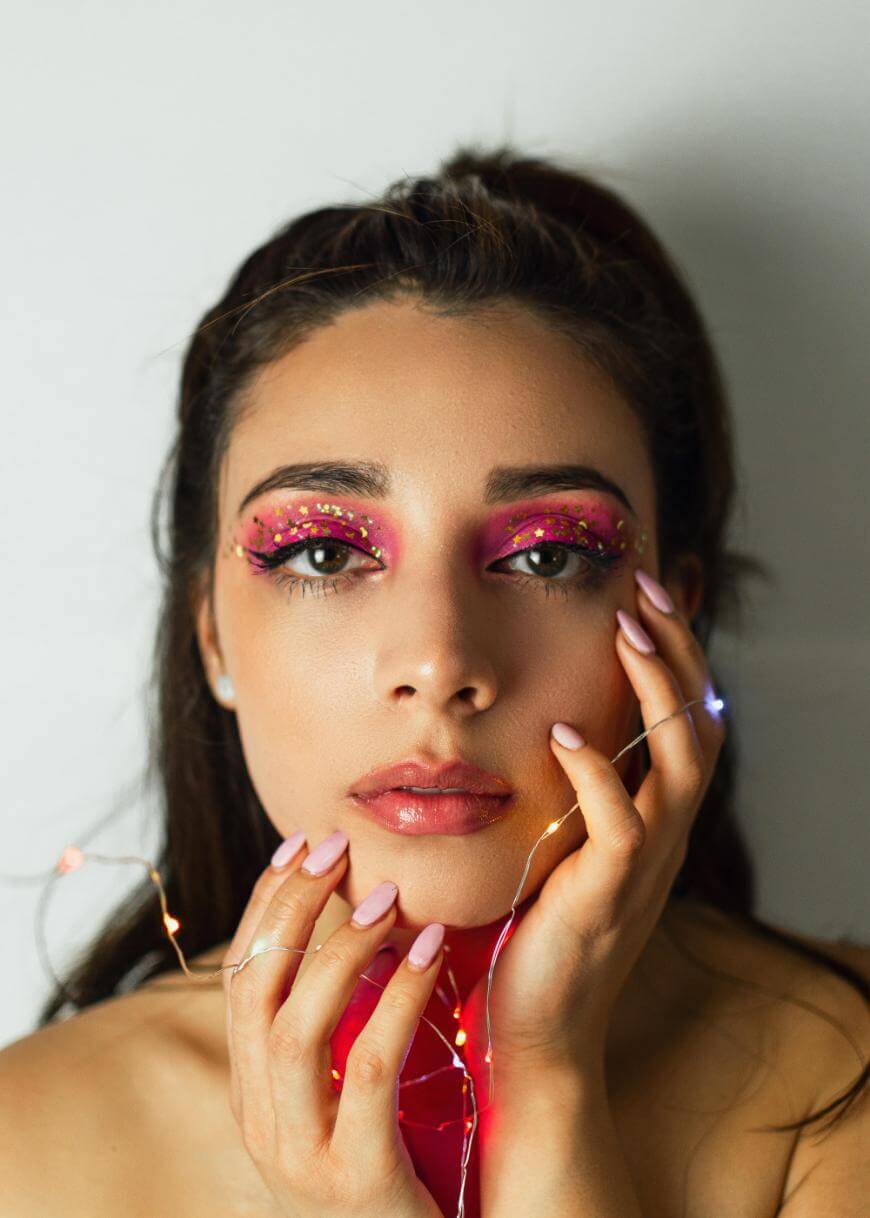 A woman with dramatic pink and glittery eye makeup gazes at the camera, holding her face with both hands. She has long, manicured nails and is wrapped in colorful string lights against a plain background.