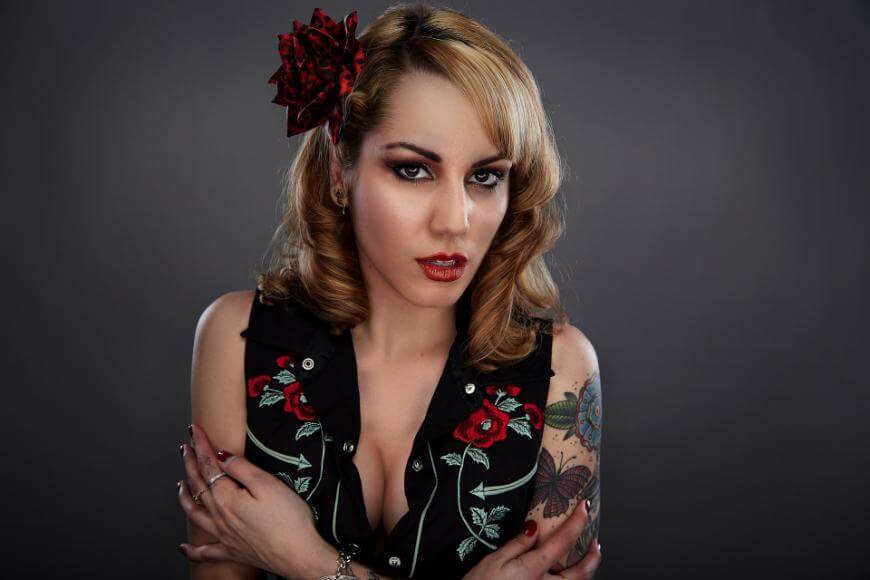 Woman with wavy blonde hair, red flower in her hair, and bold makeup, wearing a black sleeveless top with red floral embroidery, crosses her arms and looks confidently at the camera against a dark background.