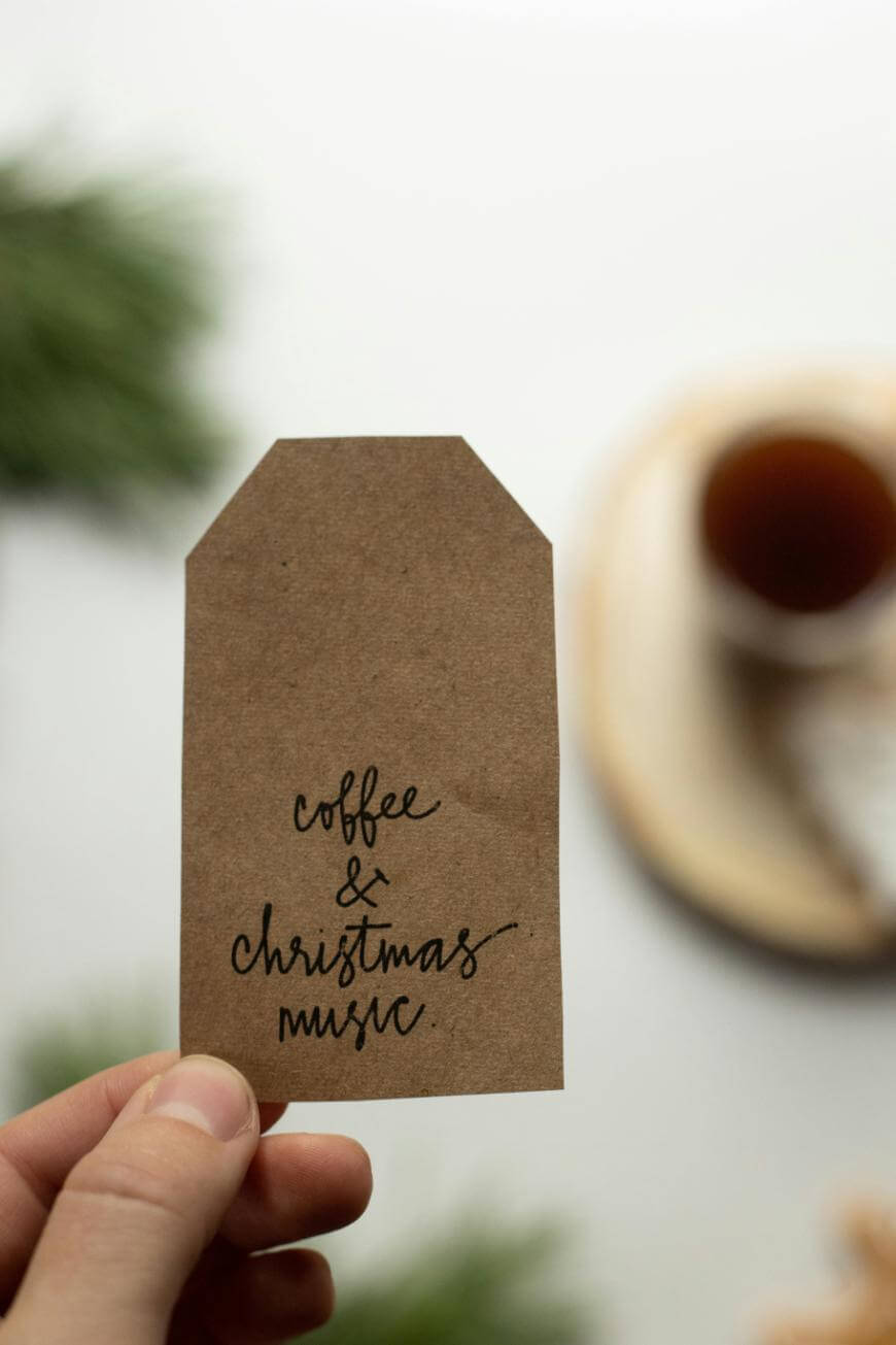 A hand holds a brown tag that reads coffee & Christmas music in cursive, with a blurred background featuring a cup of coffee and green pine branches.