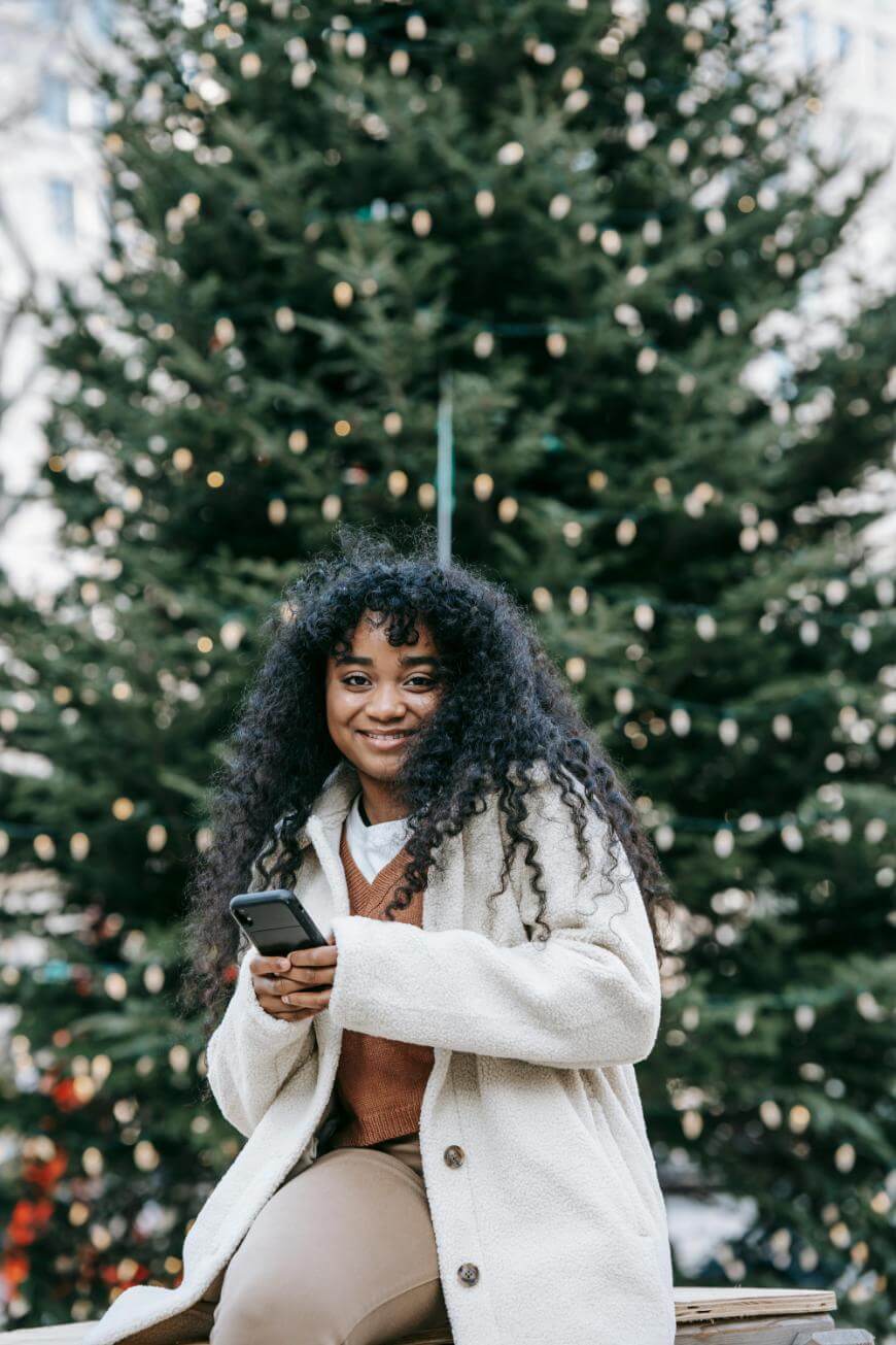 A woman with long curly hair, wearing a light coat and holding a smartphone, smiles while sitting in front of a large, decorated outdoor Christmas tree with string lights.