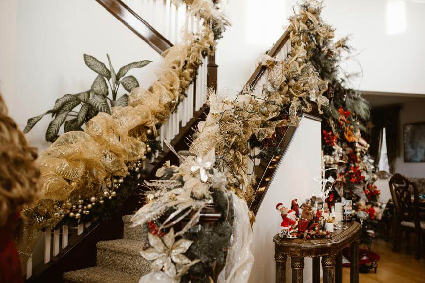 A staircase decorated for Christmas with gold mesh ribbon, ornaments, greenery, and poinsettias. Nearby, a table displays festive figurines including Santa Claus, with more holiday décor visible in the background.