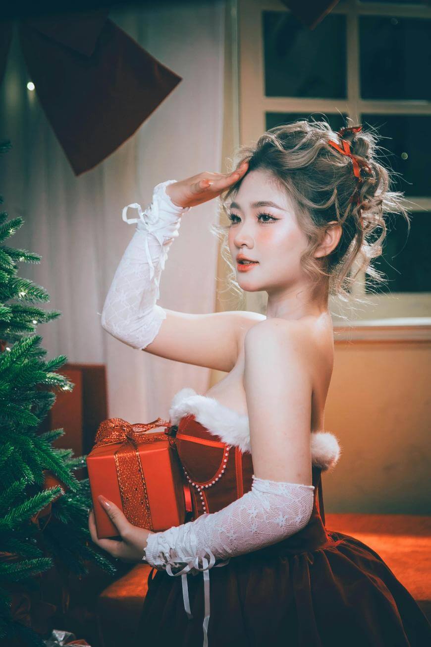 A woman in a festive red and white dress with lace gloves holds a red gift box near a decorated Christmas tree, posing with one hand raised to her forehead in a salute. Warm indoor lighting sets a holiday mood.