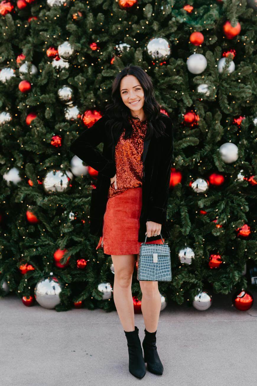 A woman stands smiling in front of a decorated Christmas tree. She wears a red top, red skirt, black jacket, black ankle boots, and holds a small handbag. The tree is adorned with red and silver ornaments.