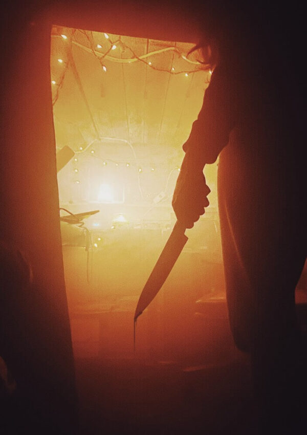 A shadowy figure holding a large knife stands in a dimly lit room with orange light and string lights in the background, creating a suspenseful and eerie atmosphere.