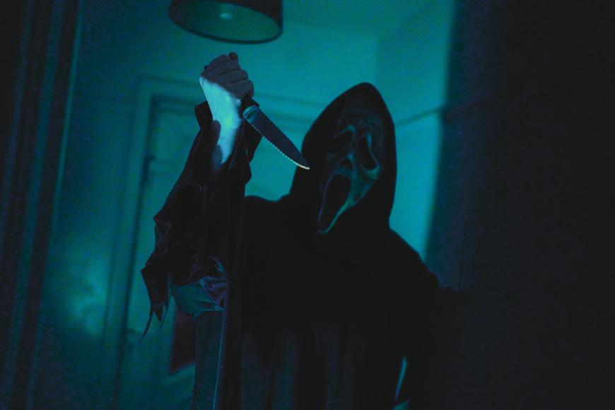 A person wearing a black hooded cloak and a white, ghost-like mask holds a large knife raised in one hand, standing in a dimly lit, eerie room.