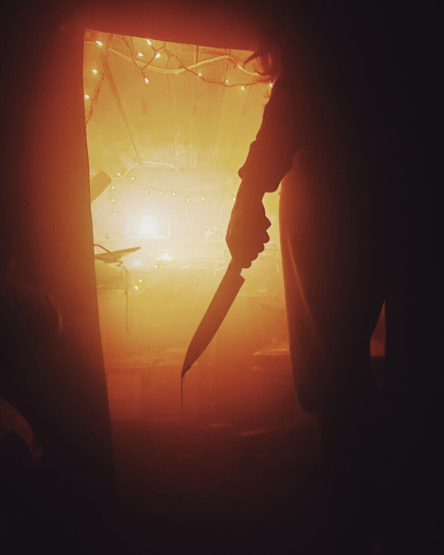 A person stands in a dimly lit, orange-tinted room holding a large knife, with their figure silhouetted in the foreground and string lights visible in the background.