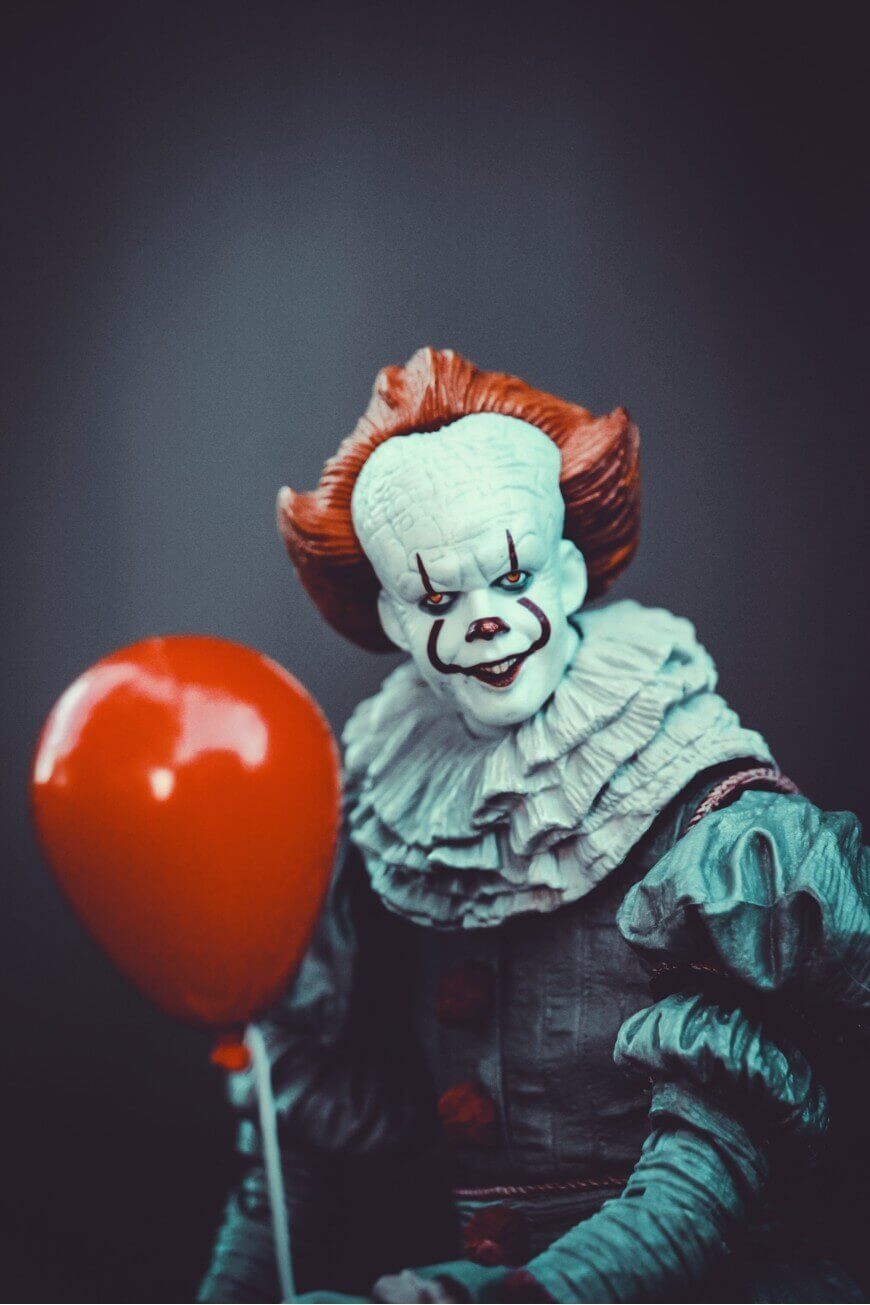 A creepy clown figurine with white makeup, red hair, and a ruffled collar holds a red balloon against a dark background.