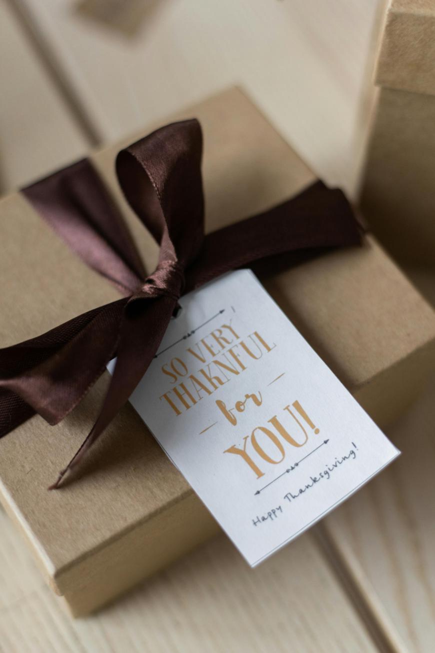 A brown gift box tied with a dark brown ribbon and a tag that reads “So very thankful for you! Happy Thanksgiving!” sits on a light wooden surface.