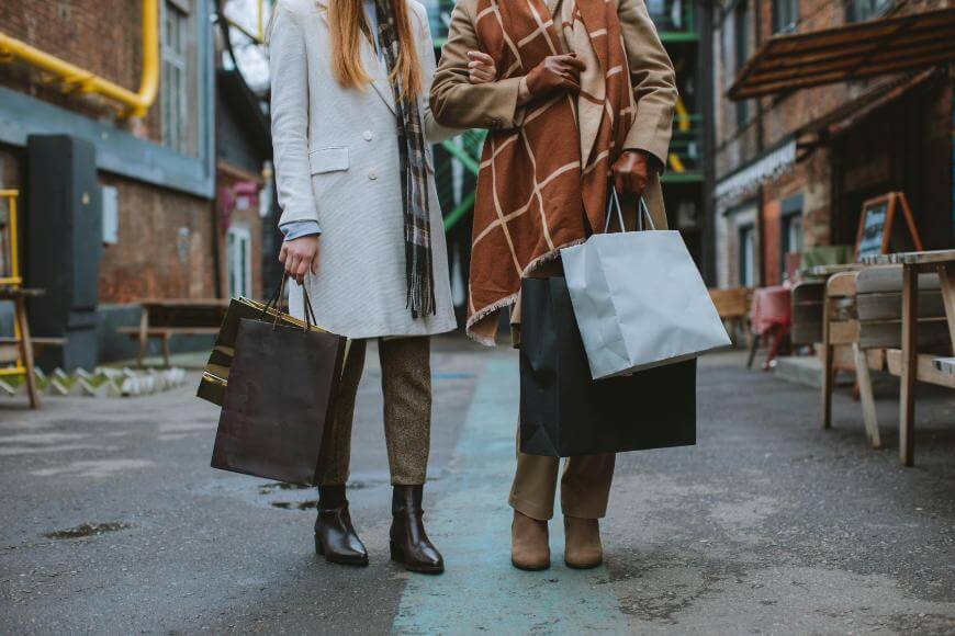 Two women, seen from the shoulders down, walk arm in arm outdoors while holding shopping bags. They wear stylish coats and boots, with one woman draped in a brown checkered scarf. The street is lined with buildings and benches.