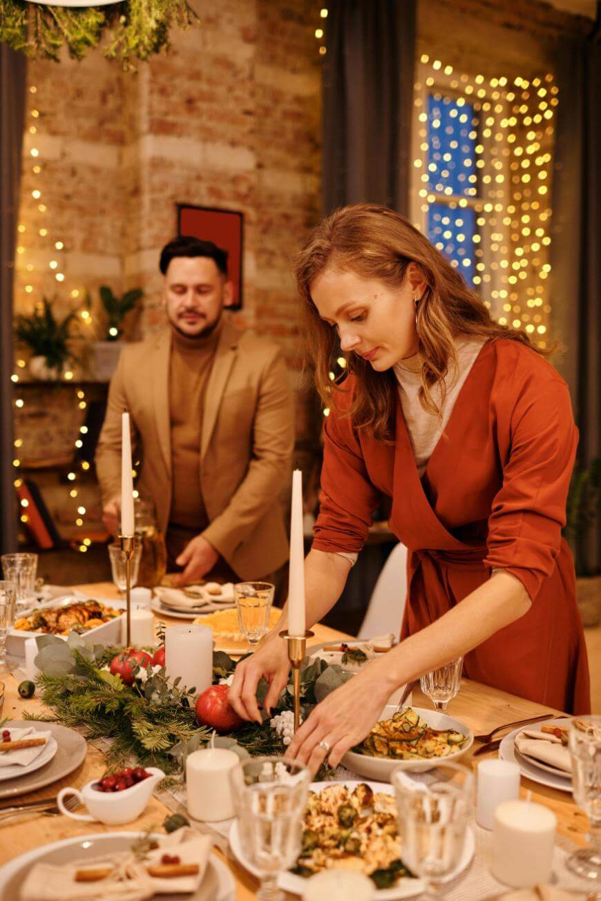 A woman in a rust-colored dress arranges a festive dinner table with candles and greenery, while a man in a brown suit stands nearby. Warm lights and holiday decorations fill the cozy, brick-walled room.