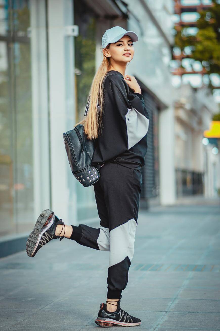 A young woman in a black and gray tracksuit, black sneakers, and a light blue cap poses on a city sidewalk with one foot raised and a black backpack over her shoulder.