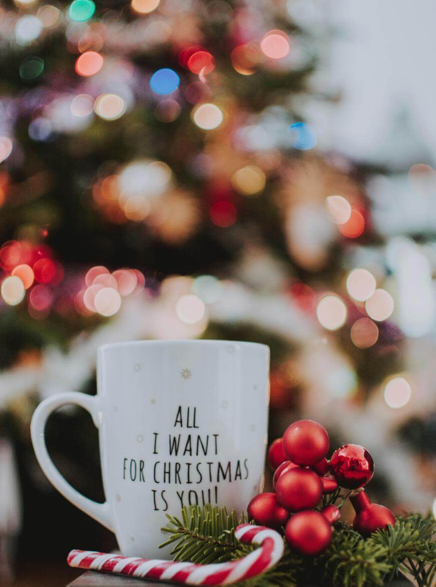 A white mug with the text All I want for Christmas is you! sits in front of a decorated Christmas tree with colorful lights. A candy cane and red ornaments are placed beside the mug.