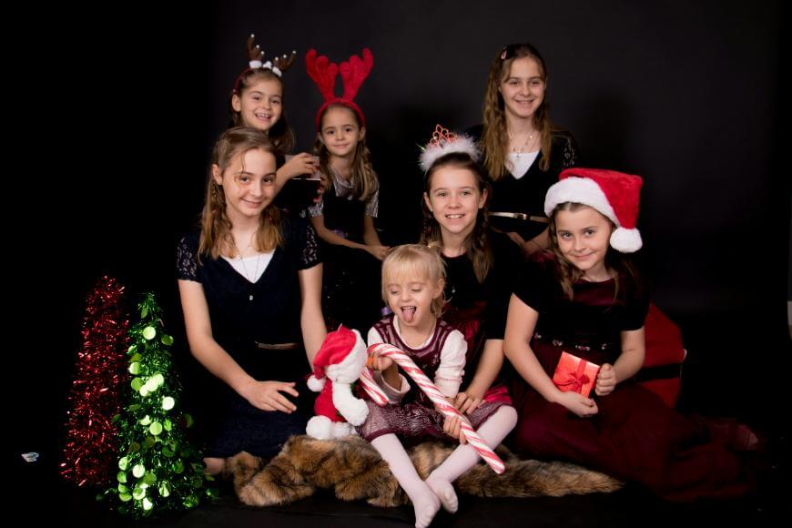Six young girls in festive outfits pose against a black background. Some wear Christmas hats, antlers, or tiaras. A toddler in front holds a large candy cane. Tinsel and a small tree decorate the scene.