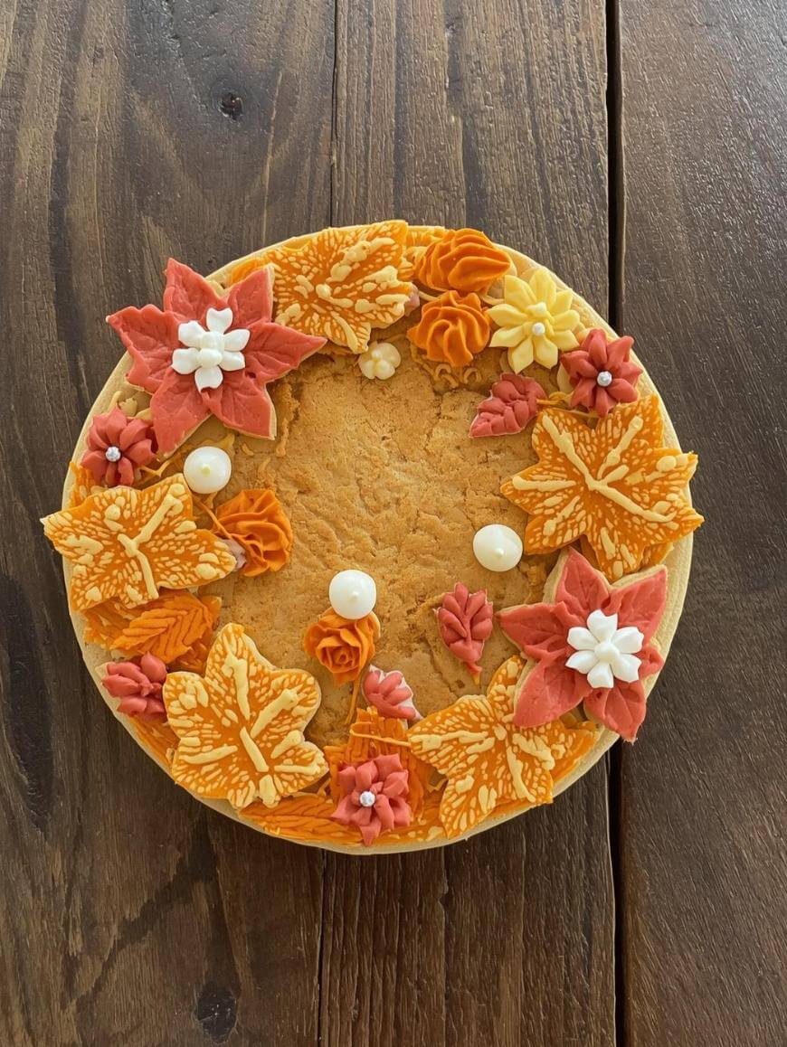 A round cake decorated with autumn-themed icing, featuring orange and yellow leaf shapes, white and orange flowers, and small white round decorations, set on a wooden surface.