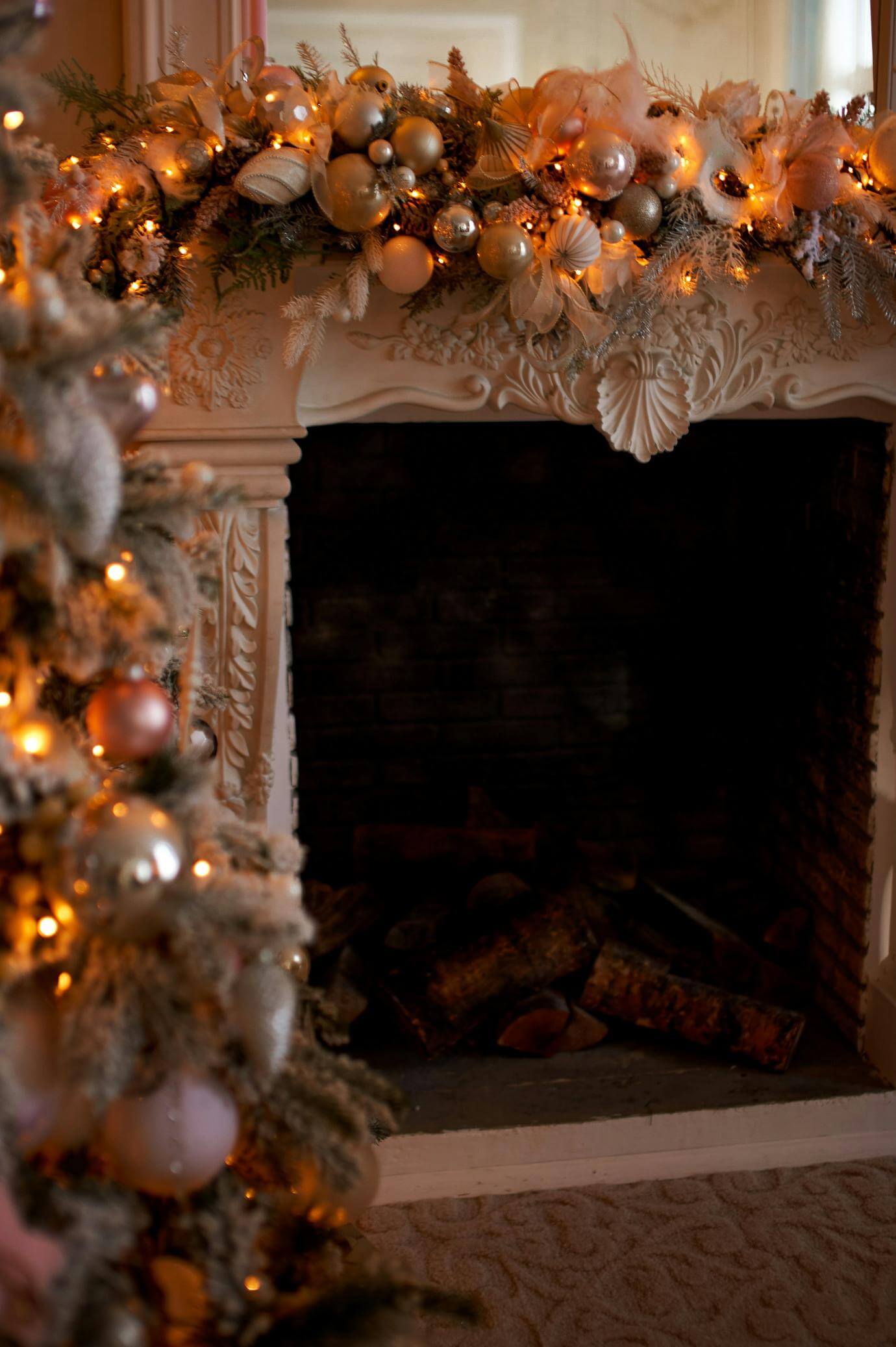 A decorated fireplace with ornaments, golden garland, and festive lights sits behind a Christmas tree adorned with similar ornaments and warm white lights. Logs are stacked in the fireplace, and the scene glows softly.