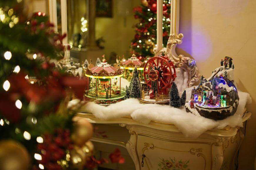 A decorative dresser topped with artificial snow displays miniature Christmas village scenes, including a carousel, Ferris wheel, and illuminated houses beside a decorated Christmas tree and a large mirror.
