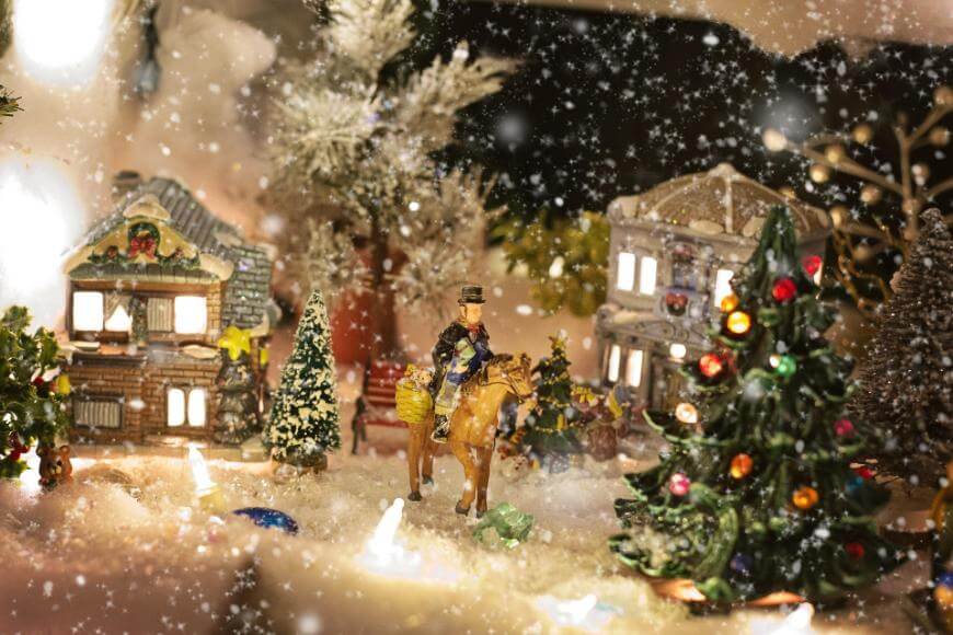 A festive miniature Christmas village scene with snow, decorated trees, small houses, and a figurine of a person riding a horse surrounded by lights and ornaments. Snowflakes appear to be falling gently.