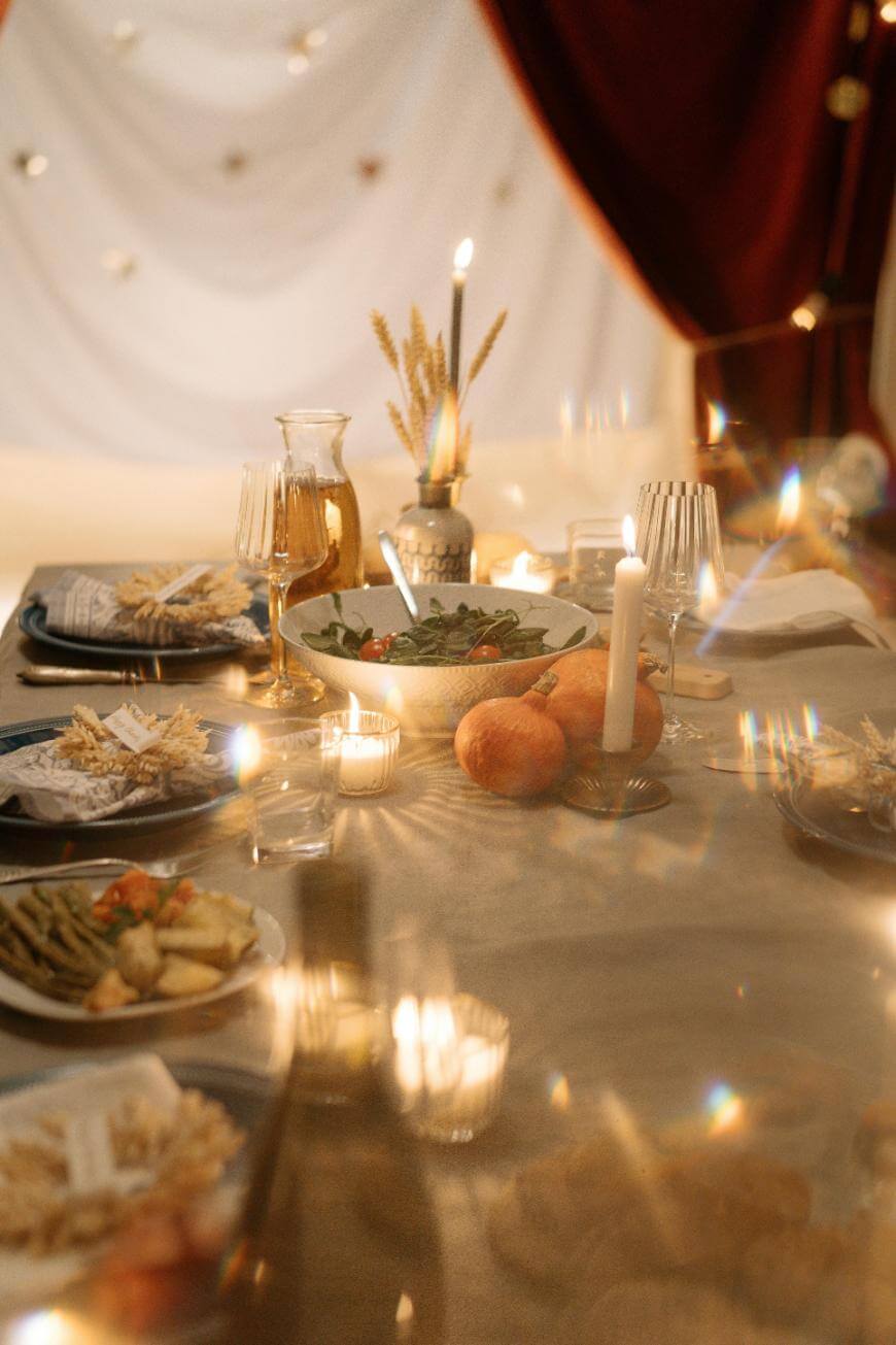 A warmly lit dining table set with plates of food, candles, glasses, a bowl of salad, and decorative gourds, with draped fabric and twinkling lights creating a cozy, festive atmosphere.