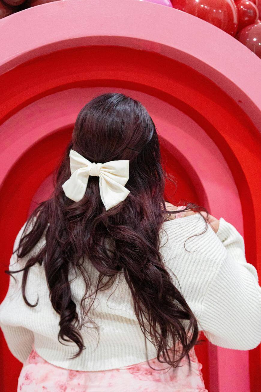 A person with long, wavy dark hair, styled with a large white bow, stands facing a red and pink arched backdrop. They wear a white sweater and a pink skirt.