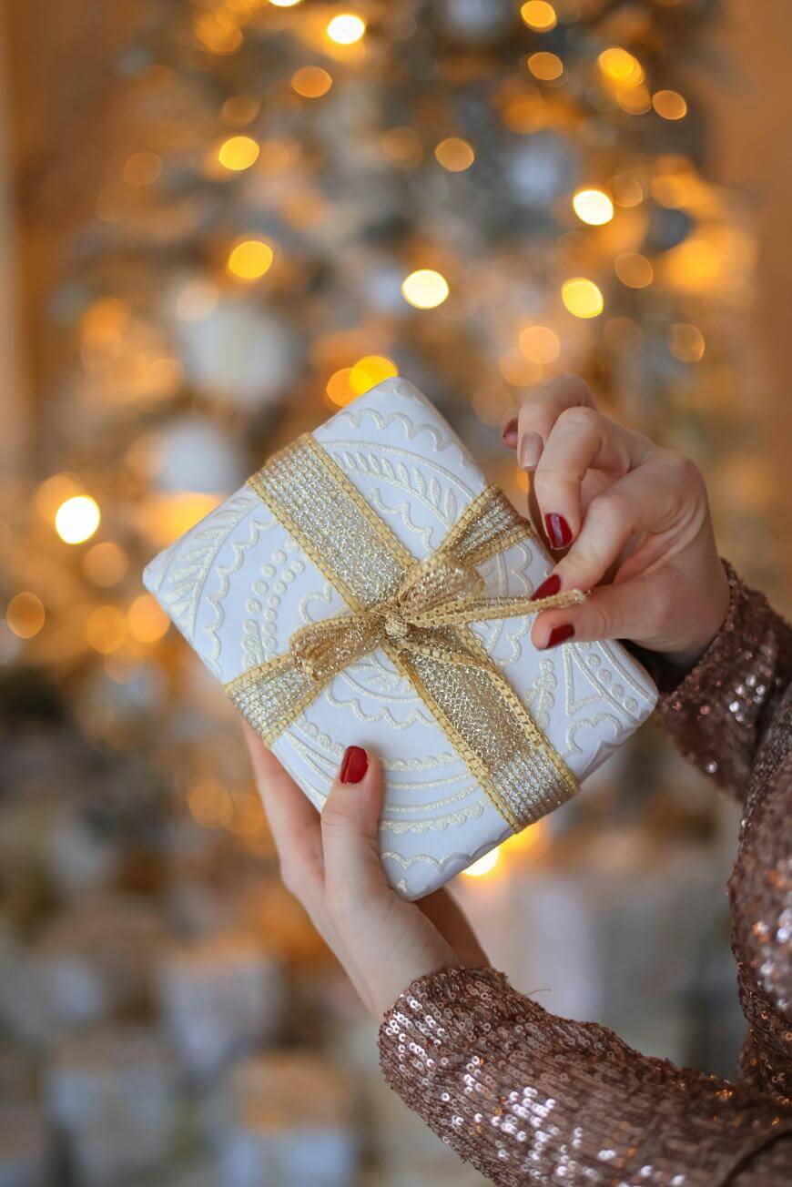 A person with red nail polish holds a white gift wrapped with a gold ribbon in front of a blurred, glowing Christmas tree. They wear a sparkly, sequined sleeve.