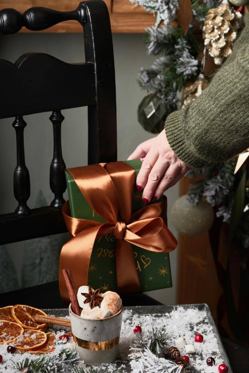 A hand places a green gift box with a brown satin ribbon on a black chair. In front, a festive table holds a cup of whipped dessert, dried orange slices, and holiday decorations dusted with snow.