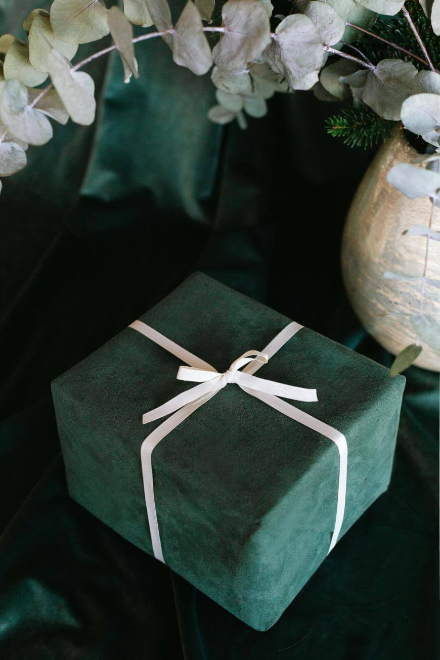 A dark green gift box tied with a white ribbon sits on a matching fabric surface next to a decorative vase with white and green foliage.