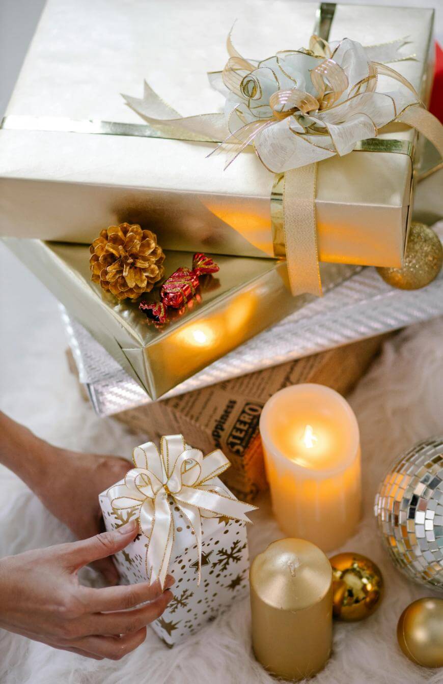 Gold and white wrapped gifts with ribbons are stacked near lit candles, a gold pine cone, Christmas ornaments, and a disco ball. A hand is reaching for a small present on a white furry surface.