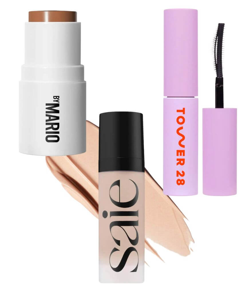 A collection of makeup products, including a brown stick foundation labeled By Mario, a liquid product labeled Saie, and a purple mascara tube labeled Tower 28, displayed on a beige smear background.