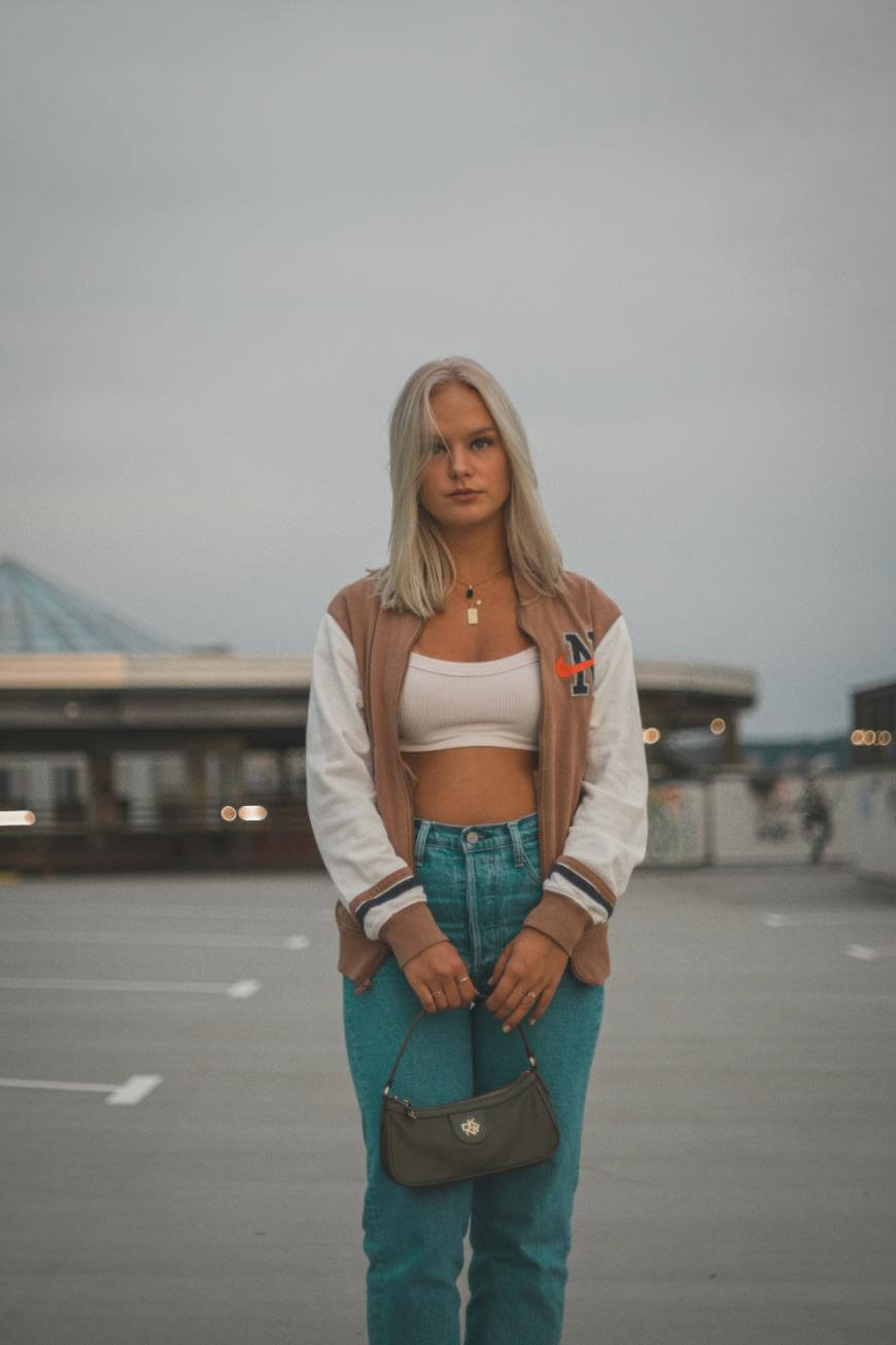 A young woman with blond hair stands on a rooftop parking lot wearing a brown and white varsity jacket, white crop top, blue jeans, and holding a small green handbag. The background is slightly blurred.