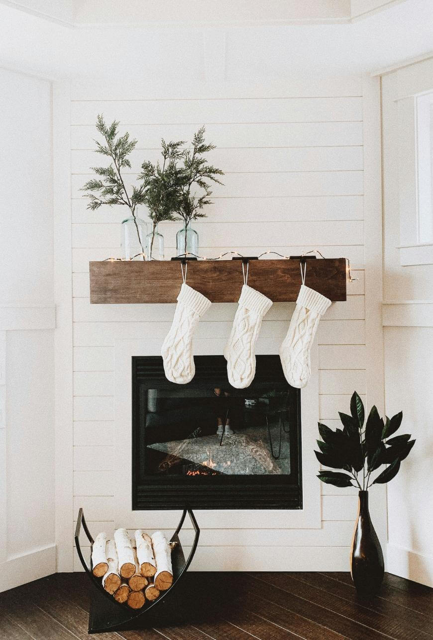 A cozy fireplace with three white knit stockings hanging from a wooden mantle, decorated with glass vases holding greenery. Firewood is stacked below, and a leafy plant in a brown vase sits on the floor beside the hearth.