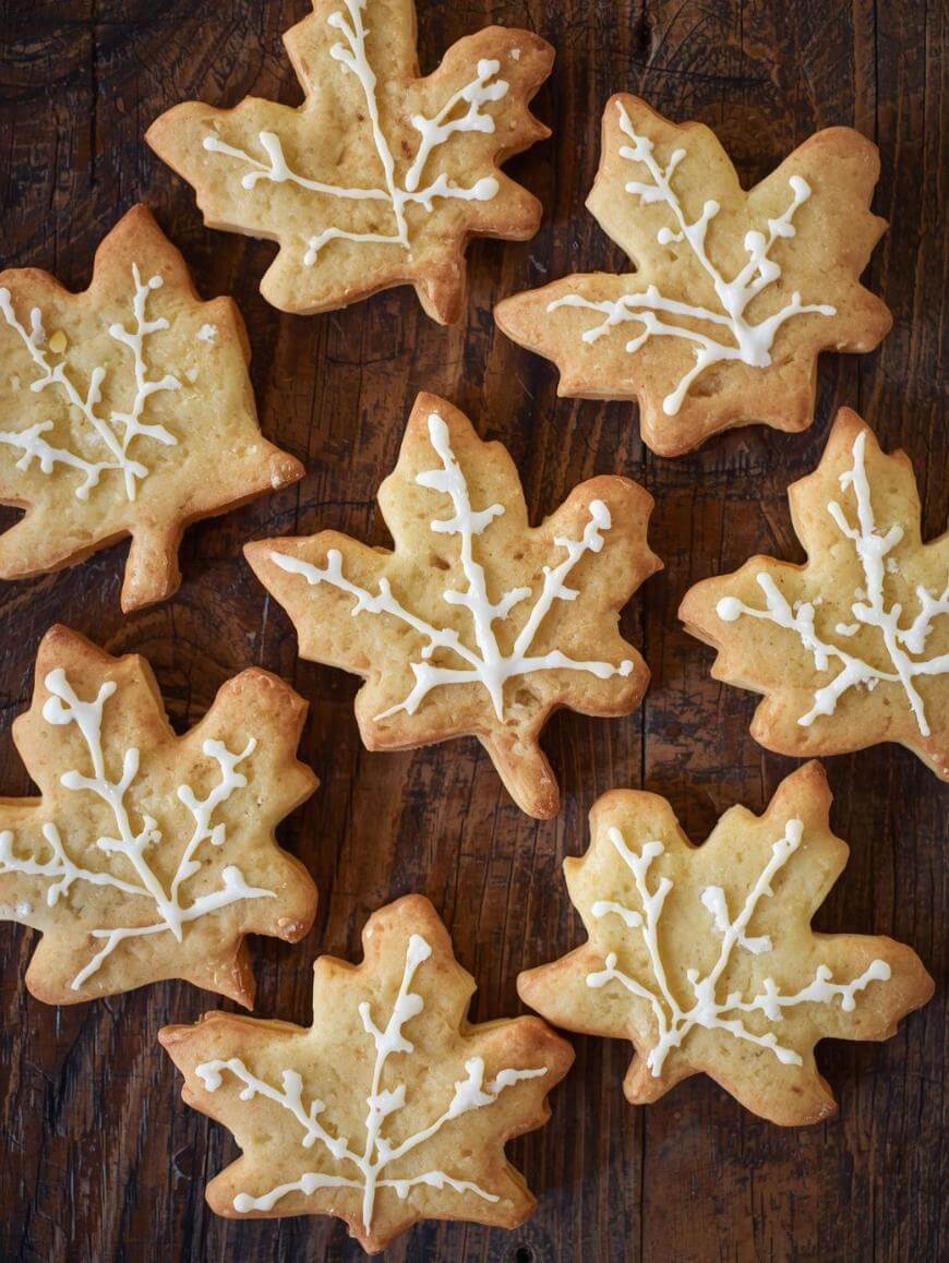 Leaf-shaped cookies with white icing designs resembling veins are arranged on a dark wooden surface. The cookies have a golden-brown color and appear homemade and festive.