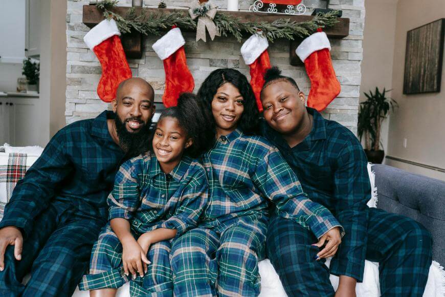 A family of four wearing matching plaid pajamas sits on a couch in front of a decorated fireplace with red stockings hanging above. They are smiling and posing for a holiday photo.