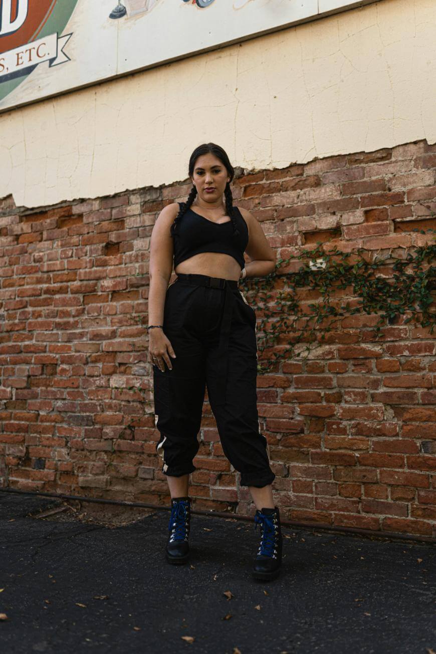 A woman with braided hair poses confidently against a weathered brick wall. She wears a black crop top, high-waisted black pants, and blue lace-up boots, with one hand on her hip. Some green vines grow on the wall nearby.