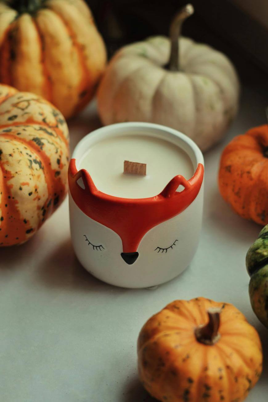 A fox-themed candle with a wooden wick sits on a white surface, surrounded by small pumpkins in shades of orange, white, and green. The candle holder has a fox face with closed eyes and red ears.