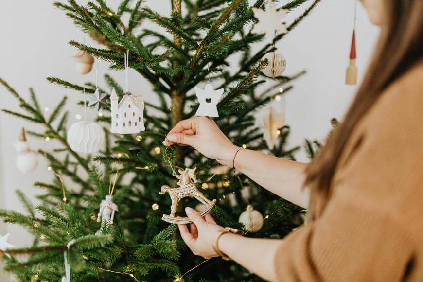 A person in a brown sweater hangs a rocking horse ornament on a decorated Christmas tree with white and wooden ornaments and twinkling lights.