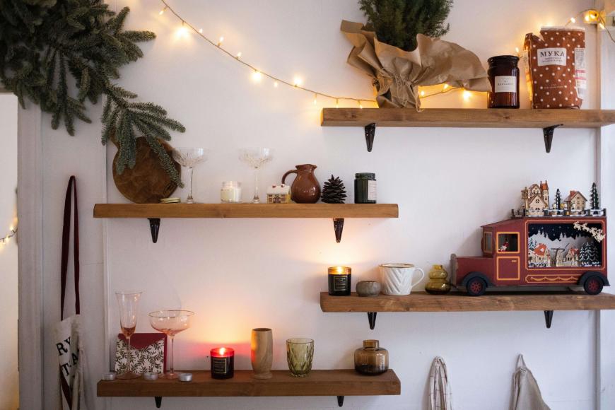 Wooden shelves on a white wall hold candles, glassware, jars, and decorative items. A string of warm fairy lights and pine branches add a cozy, festive atmosphere. A red decorative bus sits on the lower shelf.
