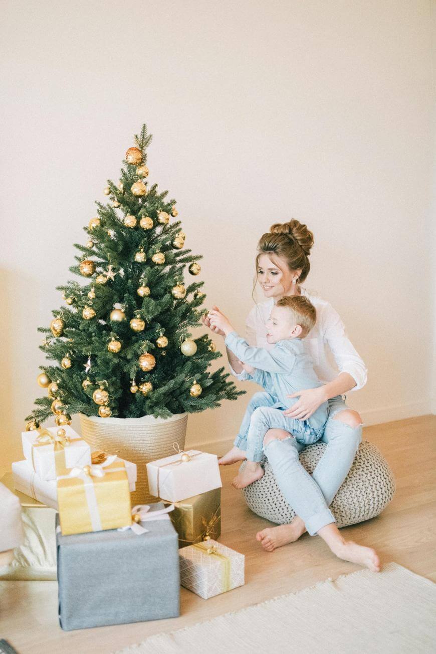 A woman and a young boy sit together by a decorated Christmas tree, hanging ornaments. Wrapped gifts with gold and white bows are stacked around the tree on a wooden floor. The scene is cozy and festive.