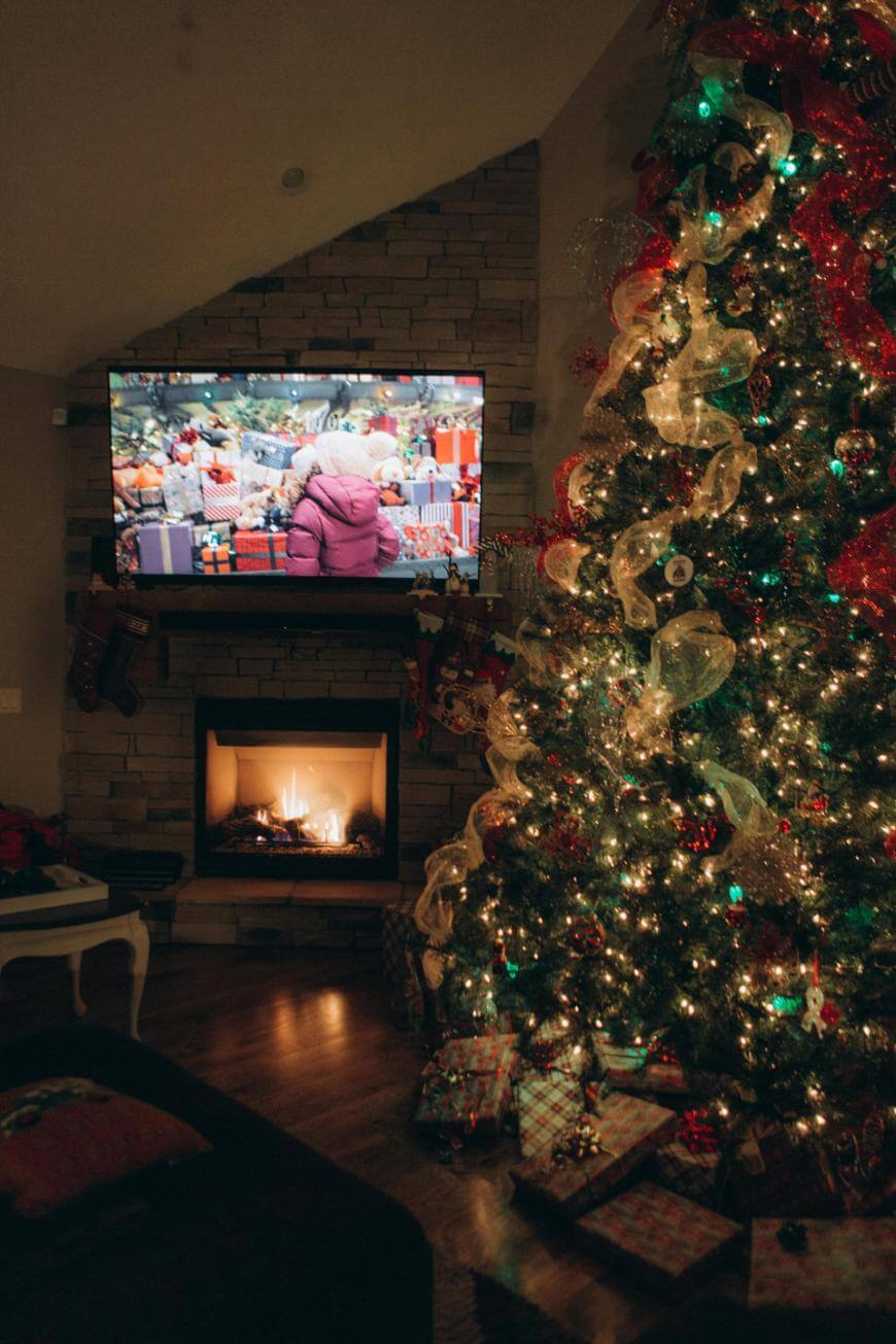 A warmly lit living room features a decorated Christmas tree with lights and ornaments, wrapped gifts underneath, a fireplace with stockings, and a TV showing a festive scene.