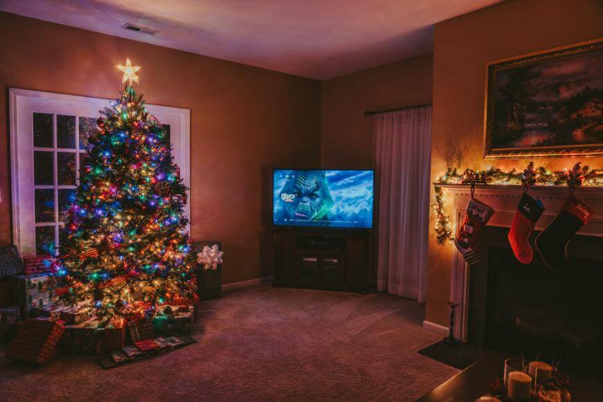 A living room decorated for Christmas with a lit, ornamented tree, wrapped presents underneath, stockings hanging on the fireplace, and a TV displaying a holiday movie. Warm, festive lighting fills the room.